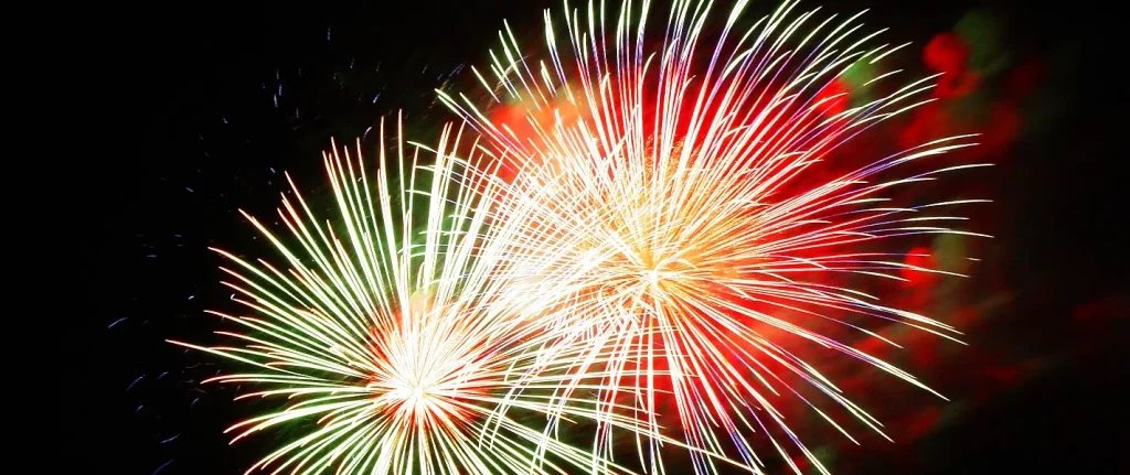 Two large overlapping firework bursts—one primarily green-to-yellow on the left and a brighter yellow-orange one on the right with red accents—against a dark sky.


